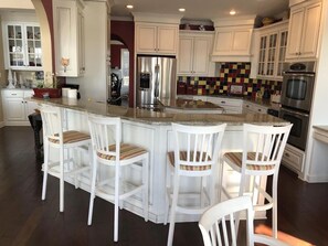 A kitchen with white cabinets, a stainless steel refrigerator, a colorful backsplash, and a bar area with white chairs.