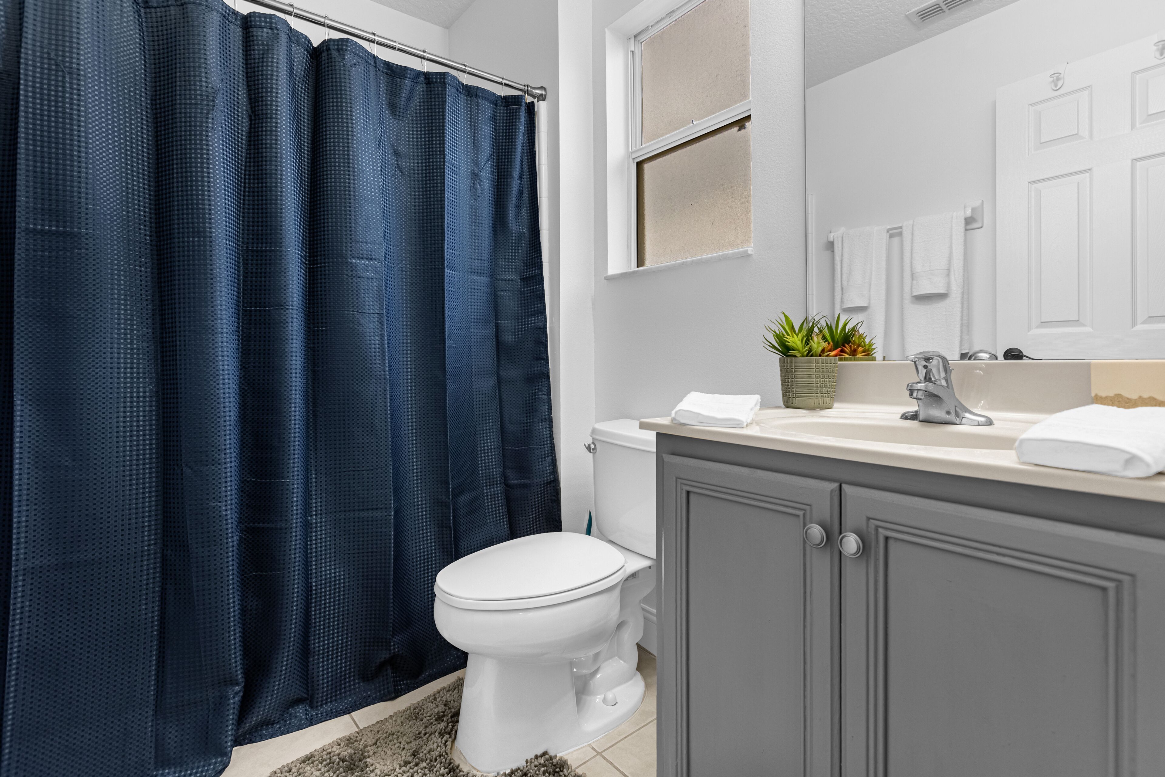Un baño con un inodoro blanco, un mueble gris, un lavabo blanco, un espejo y una cortina de ducha azul.