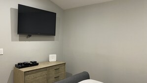 Une chambre à coucher comprenant un lit, un téléviseur fixé au mur, une commode en bois avec des tiroirs et une table de chevet avec une lampe.