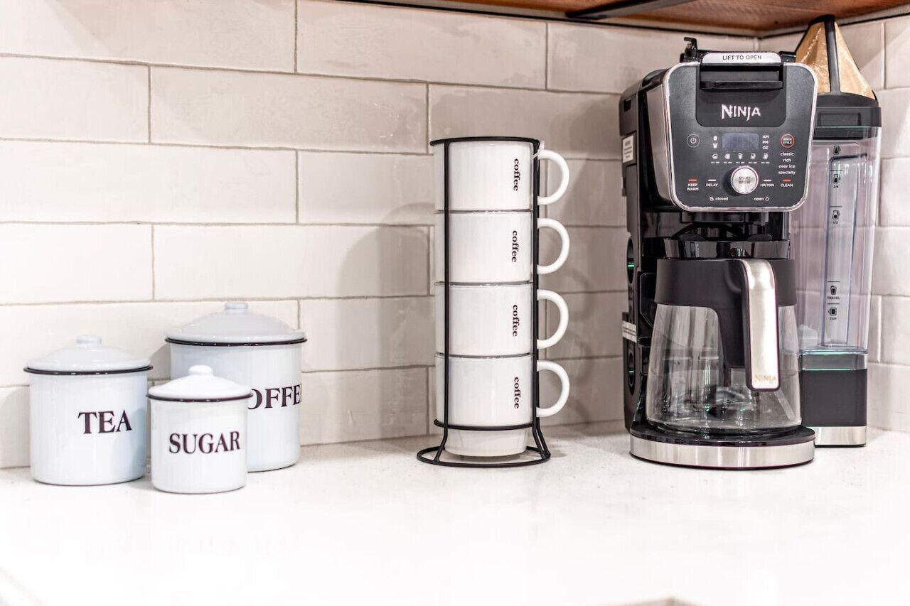 Un plan de travail de cuisine avec une machine à café, un porte-tasse et des récipients étiquetés pour le thé, le sucre et le café.