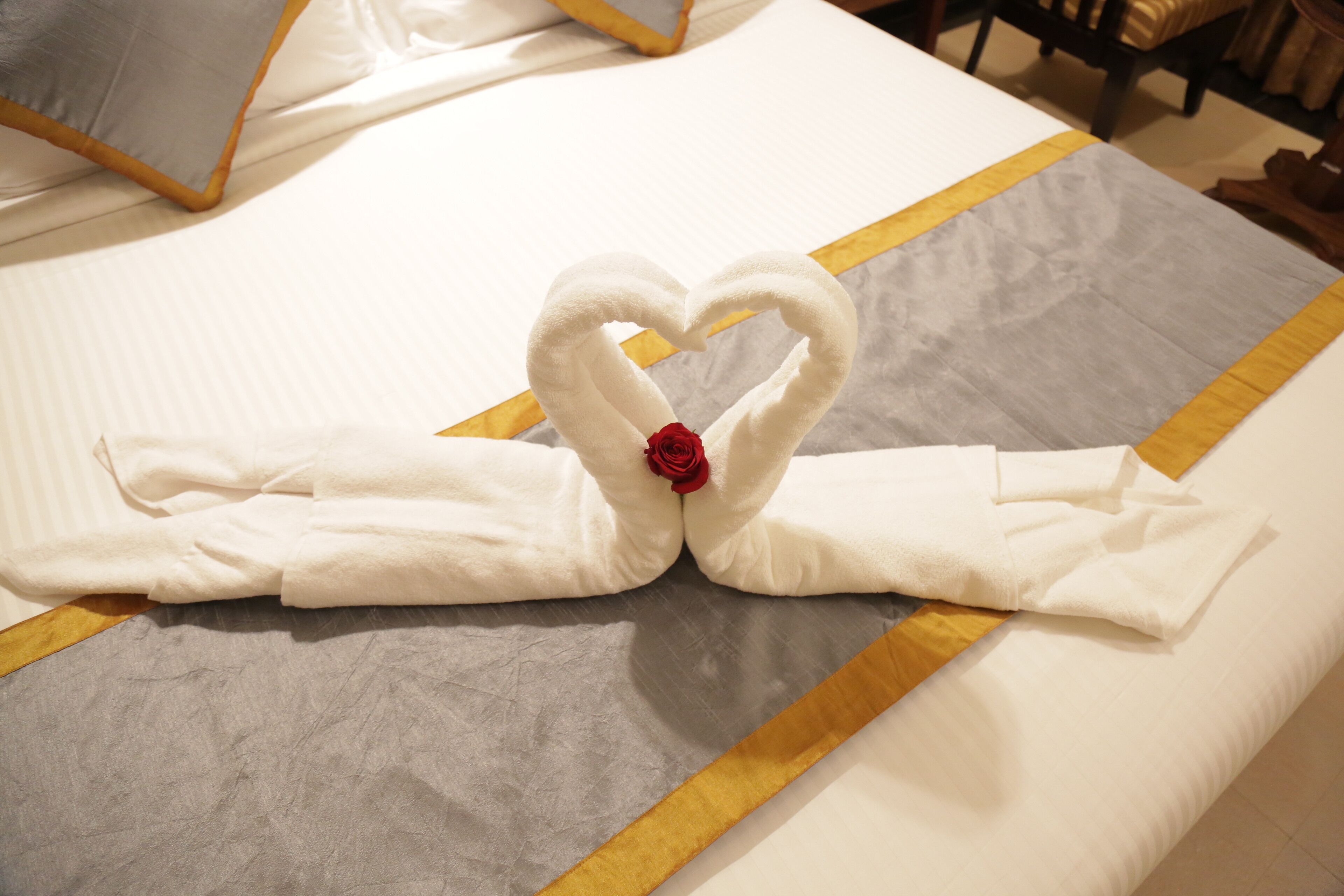 A bed with a heart-shaped towel arrangement and a red rose.