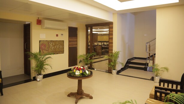A lobby with a round wooden table with a floral centerpiece, potted plants, a staircase, and a wall-mounted air conditioning unit. - Rangalaya Royal (Katpadi)