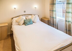 A neatly made bed with white linens and multiple pillows, two bedside tables, a radiator, and a window with curtains.