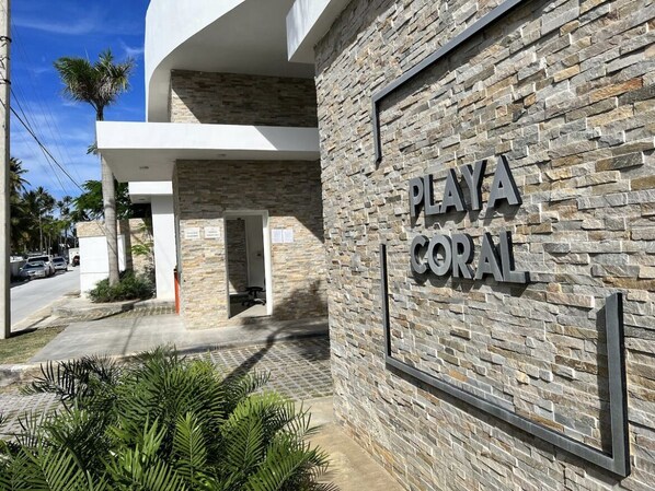 A building facade with a stone-textured wall and a sign that reads "PLAYA CORAL." - Playa Coral Condo in Paradise F22 (Punta Cana)