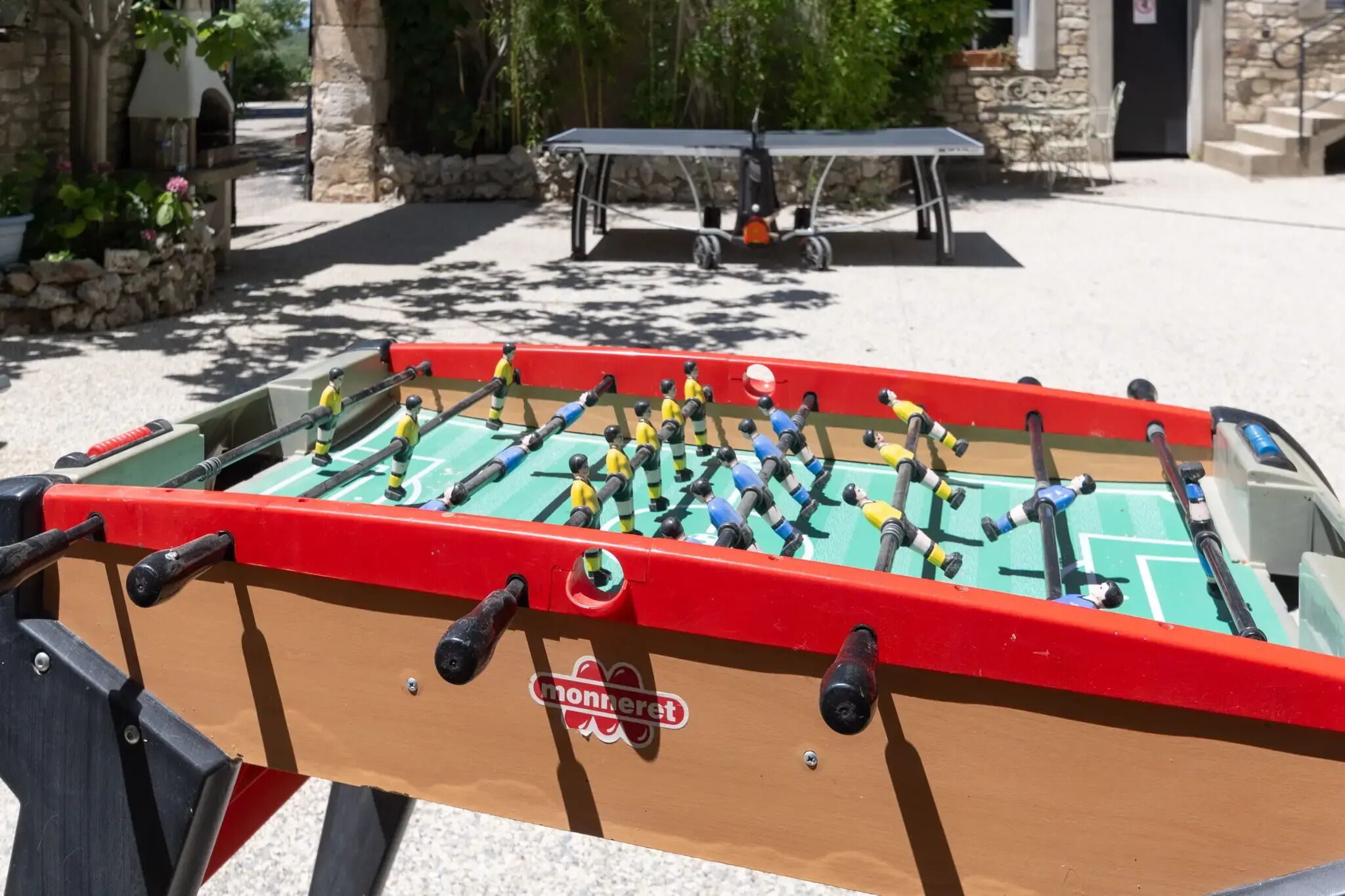 Une table de baby-foot avec un plateau rouge et vert, entourée de sièges extérieurs et de verdure.