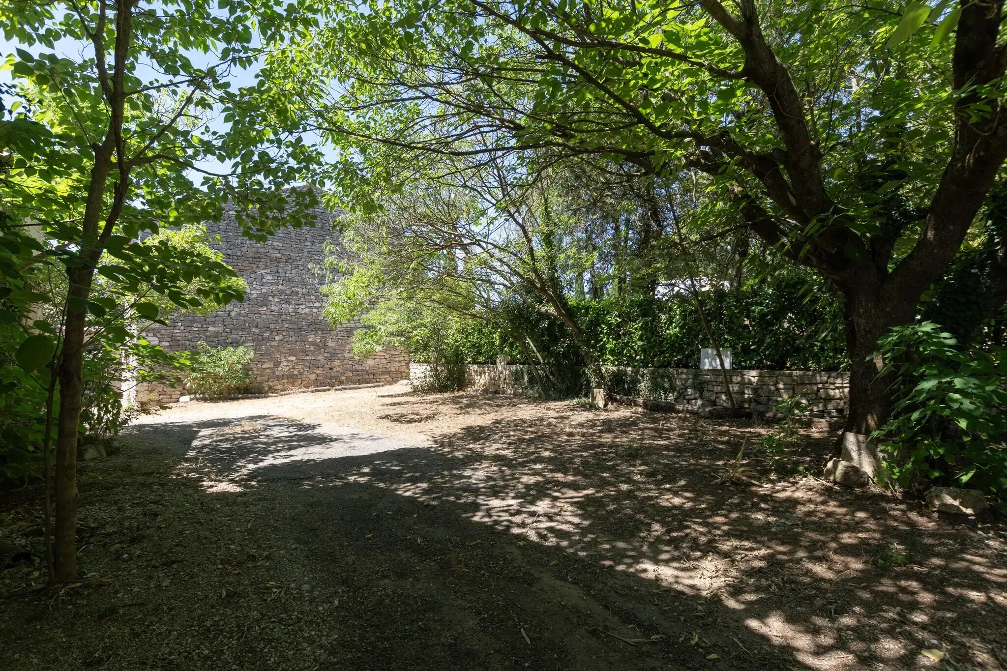 Un sentiero sterrato conduce a un muro in pietra, immerso nel verde e alberi.
