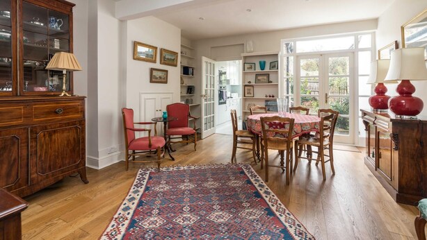 A spacious dining area with a wooden table and chairs, a patterned rug, and a view of the kitchen through an open door. - Charming 2BD Garden House - Hammersmith (London)