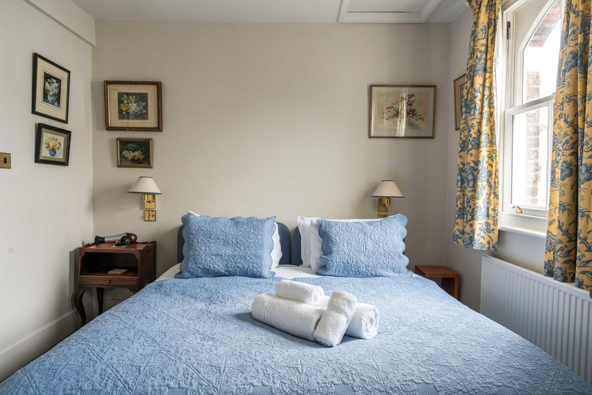 A neatly made bed with blue bedding, two bedside lamps, and a window with floral curtains.
