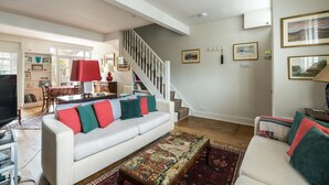 A spacious living room with a white sofa, a patterned ottoman, and a staircase leading to another level. - Charming 2BD Garden House - Hammersmith (London)