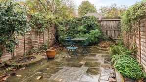 A stone patio with a table and chairs, surrounded by a wooden fence and various plants. - Charming 2BD Garden House - Hammersmith (London)