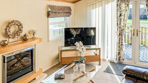 A living room with a fireplace, a television, a glass coffee table, a sofa, and a view of the outdoors. - Borwick Heights 10 is ideal for a relaxing getaway. (Carnforth)