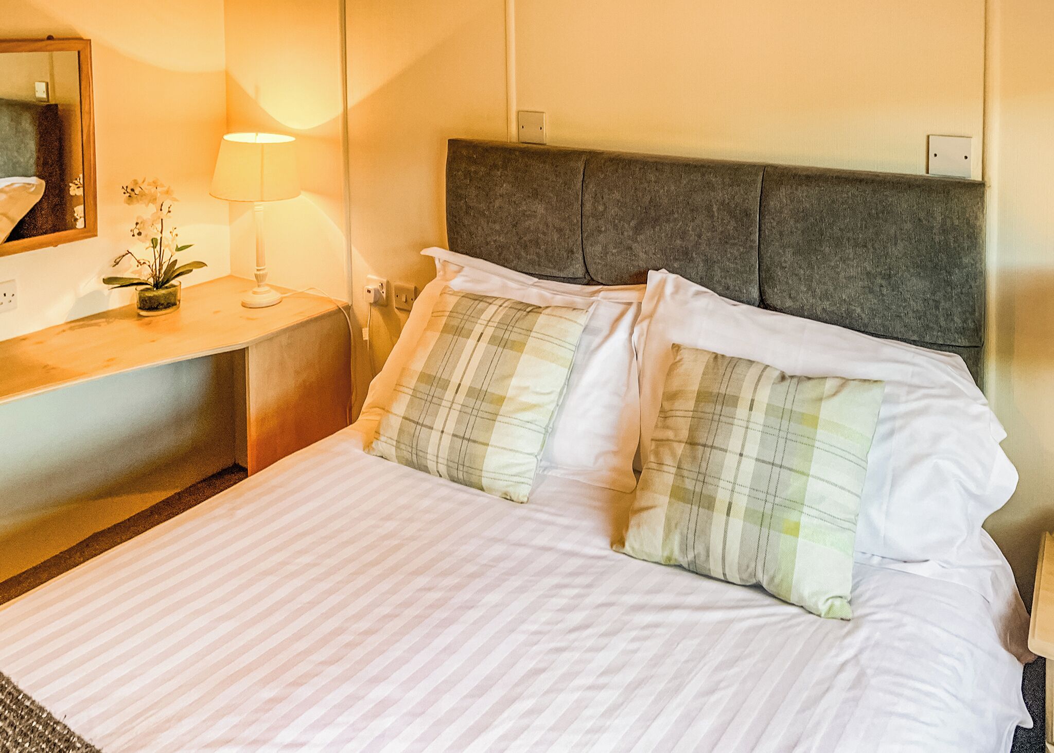 A neatly made bed with white linens and plaid pillows, a bedside table with a lamp, and a mirror on the wall.