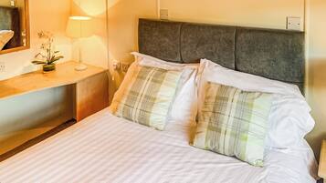 A neatly made bed with white linens and plaid pillows, a bedside table with a lamp, and a mirror on the wall.