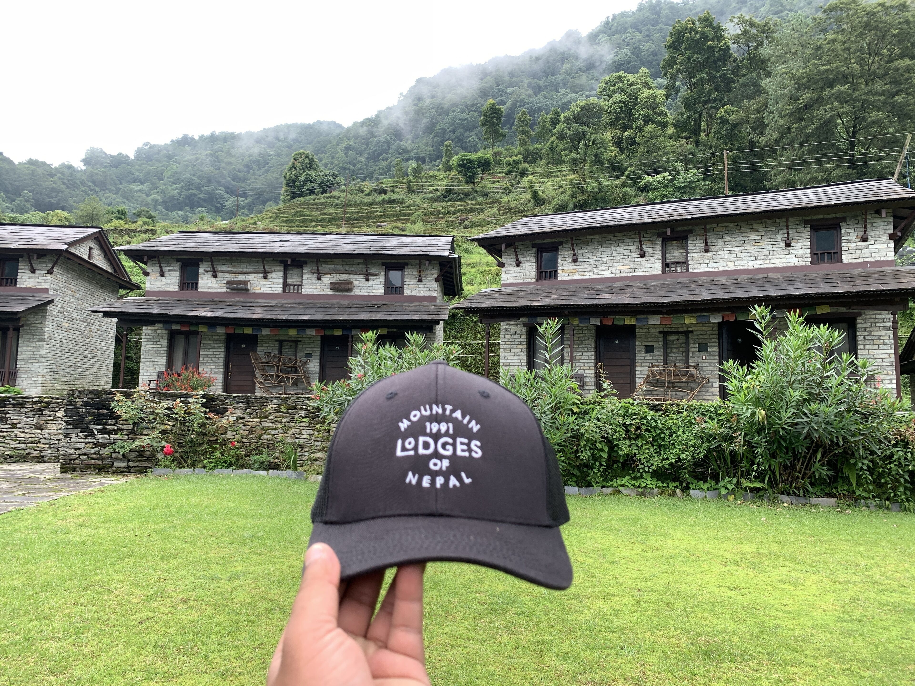 A hand holding a black cap with "Mountains of Nepal" text, in front of a stone building with a garden and a mountainous backdrop.