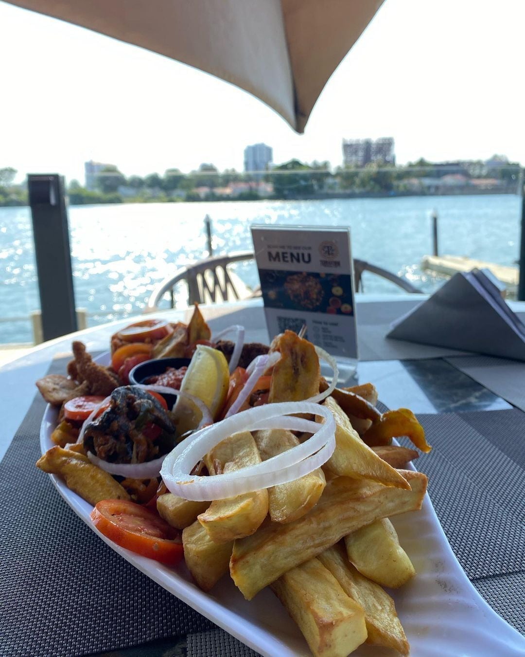 Een bord eten met friet, uien en een menukaart op een tafel aan het water.