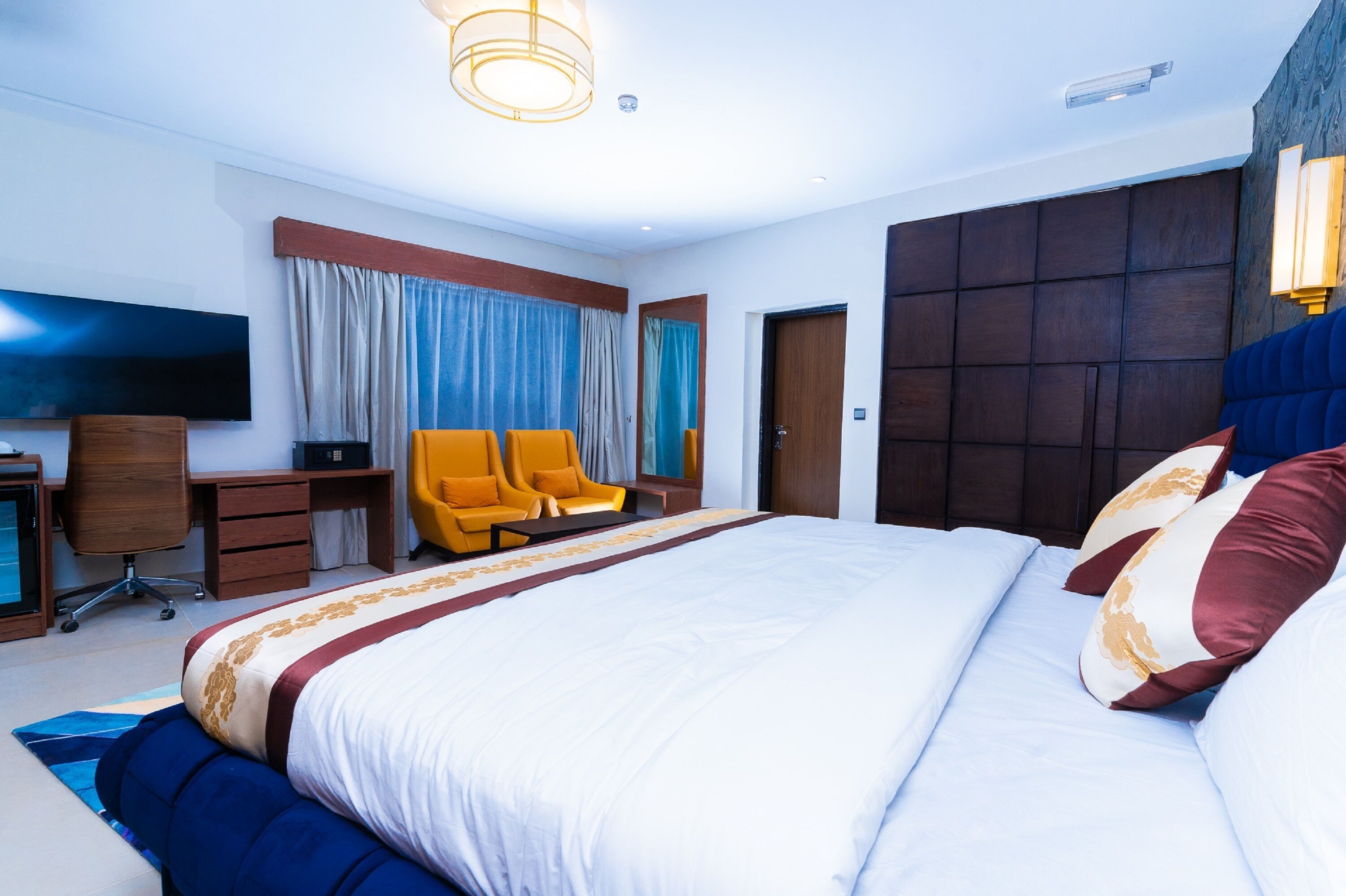 A modern hotel room with a large bed, a desk with a chair, a television, and a wardrobe.