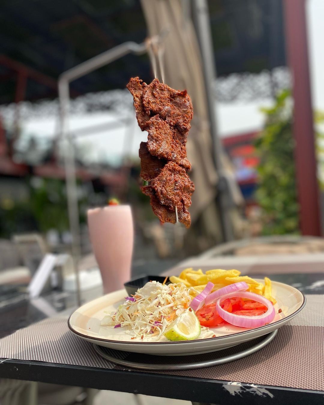 Un plato con un pincho de carne, ensalada y papas fritas sobre una mesa al aire libre.
