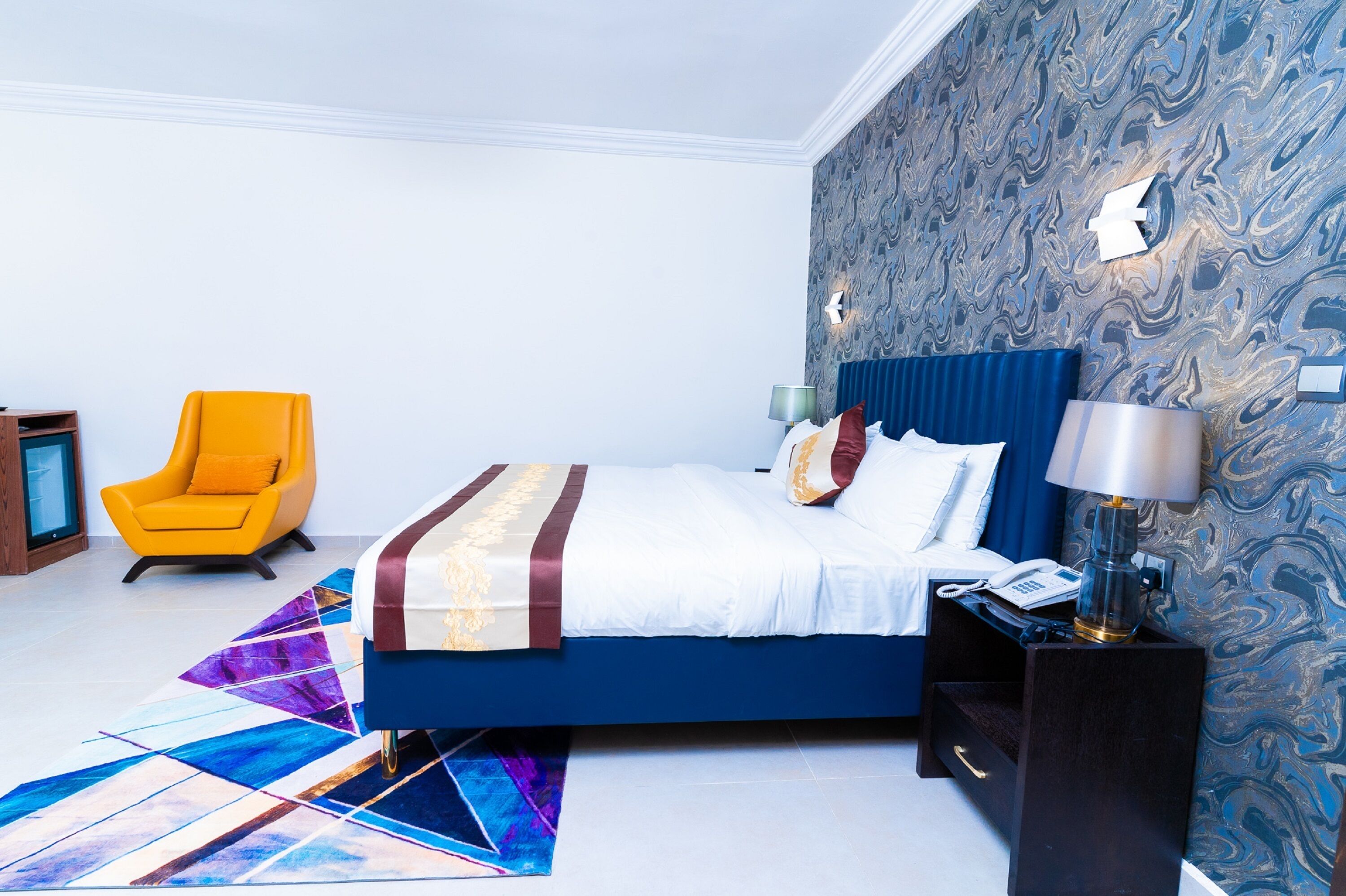 A modern bedroom with a blue bed, a yellow armchair, and a patterned wallpaper.