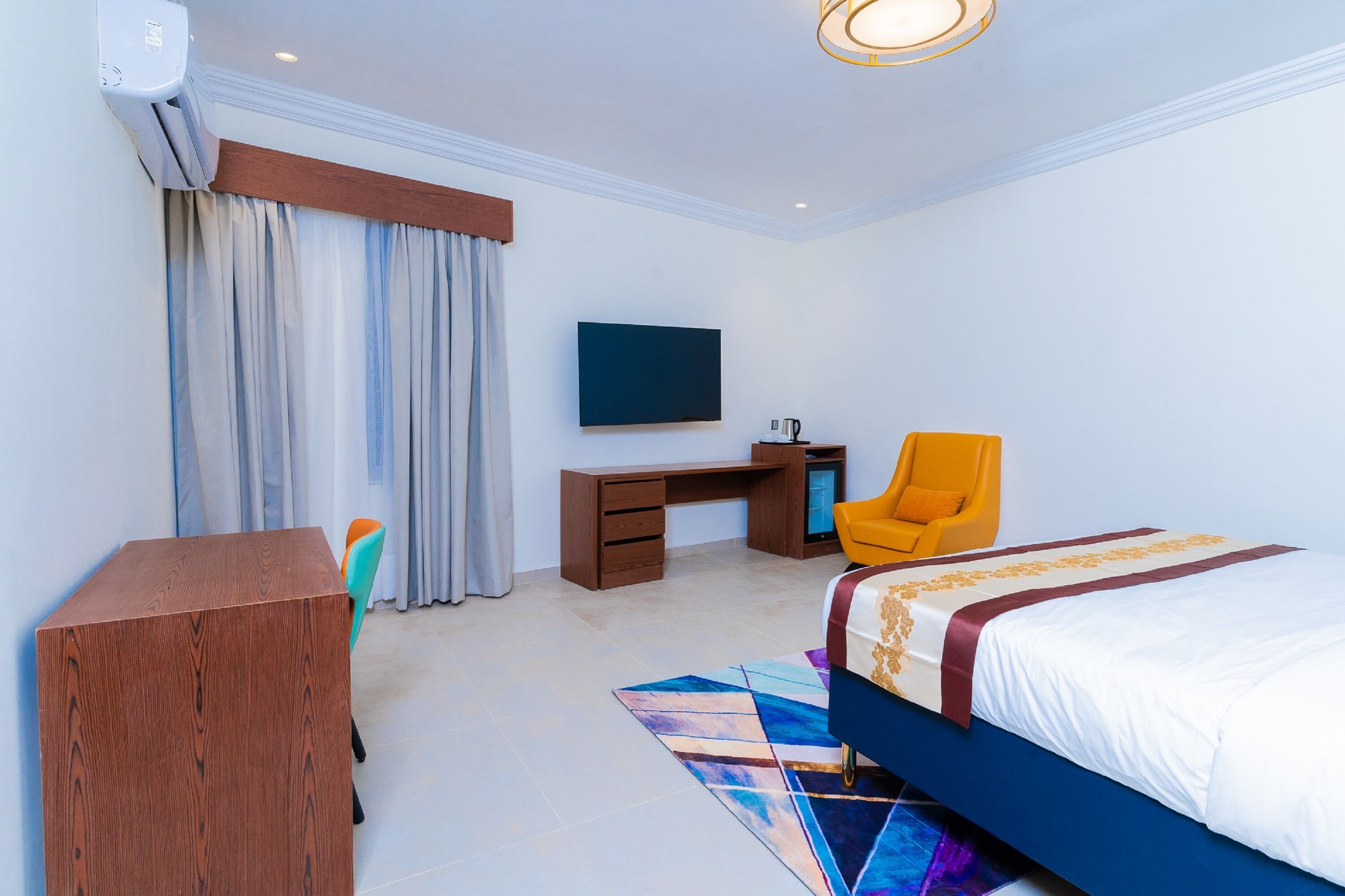 A hotel room with a bed, a desk, a chair, a TV, and an air conditioning unit.