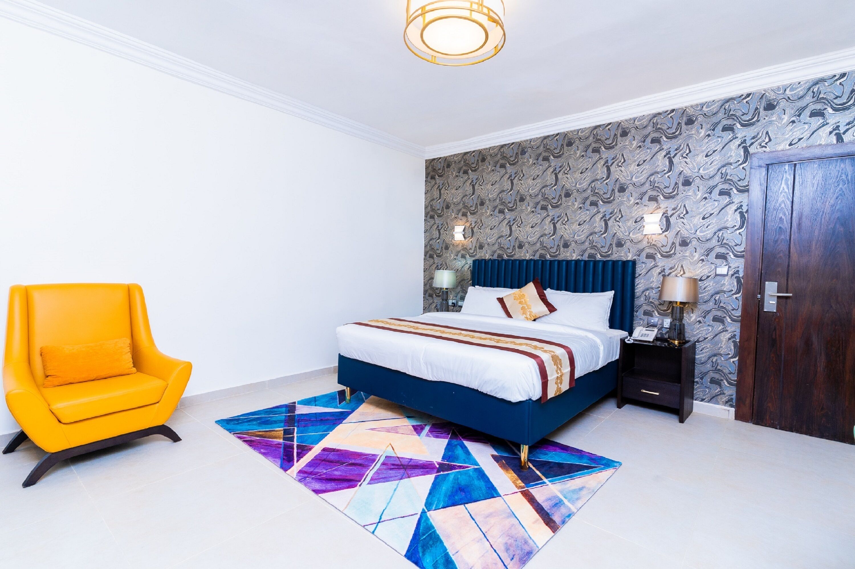 Un dormitorio con cama, mesita de noche, lámpara, silla y alfombra estampada.