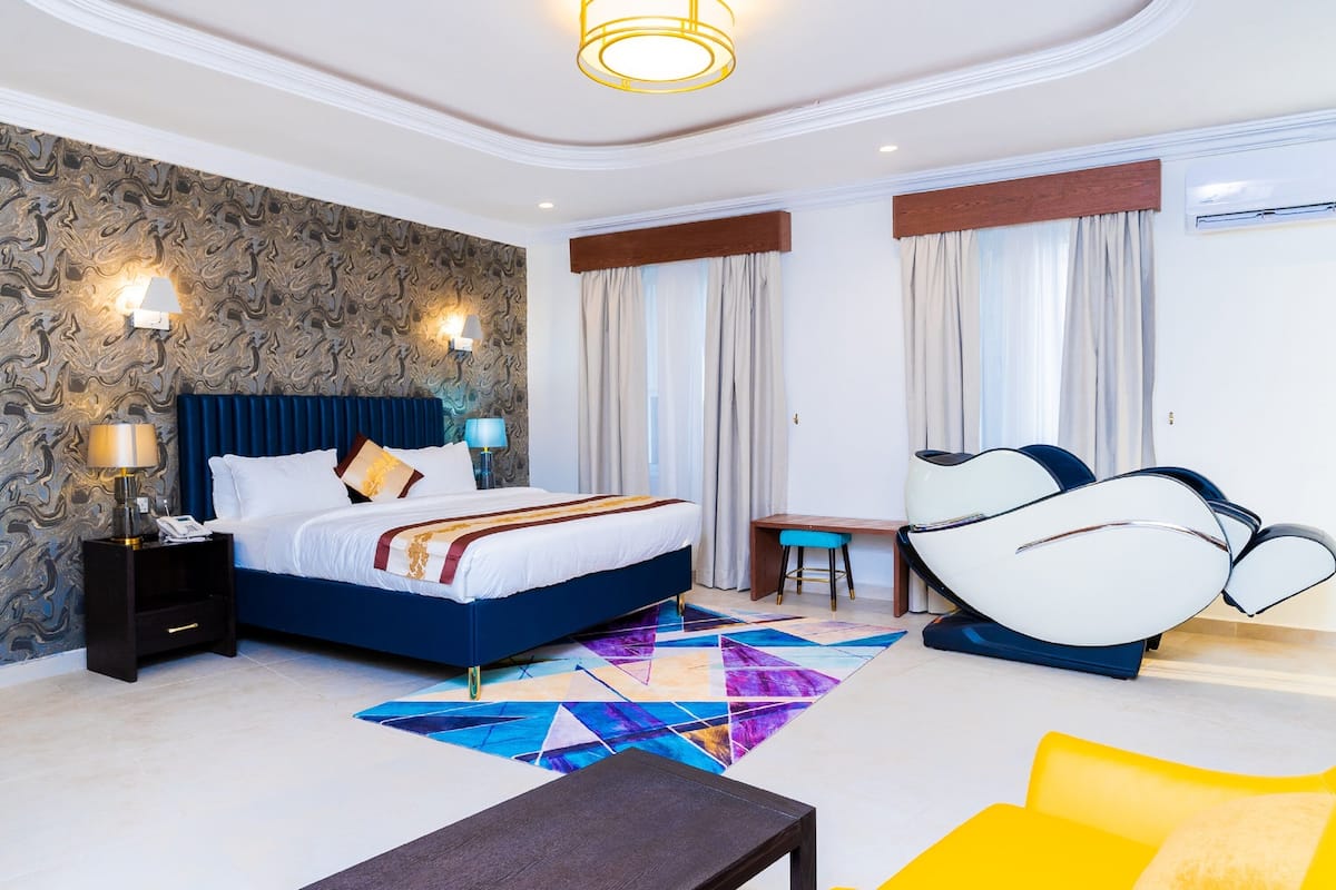 A modern hotel room with a large bed, a desk, a massage chair, and a colorful geometric rug.
