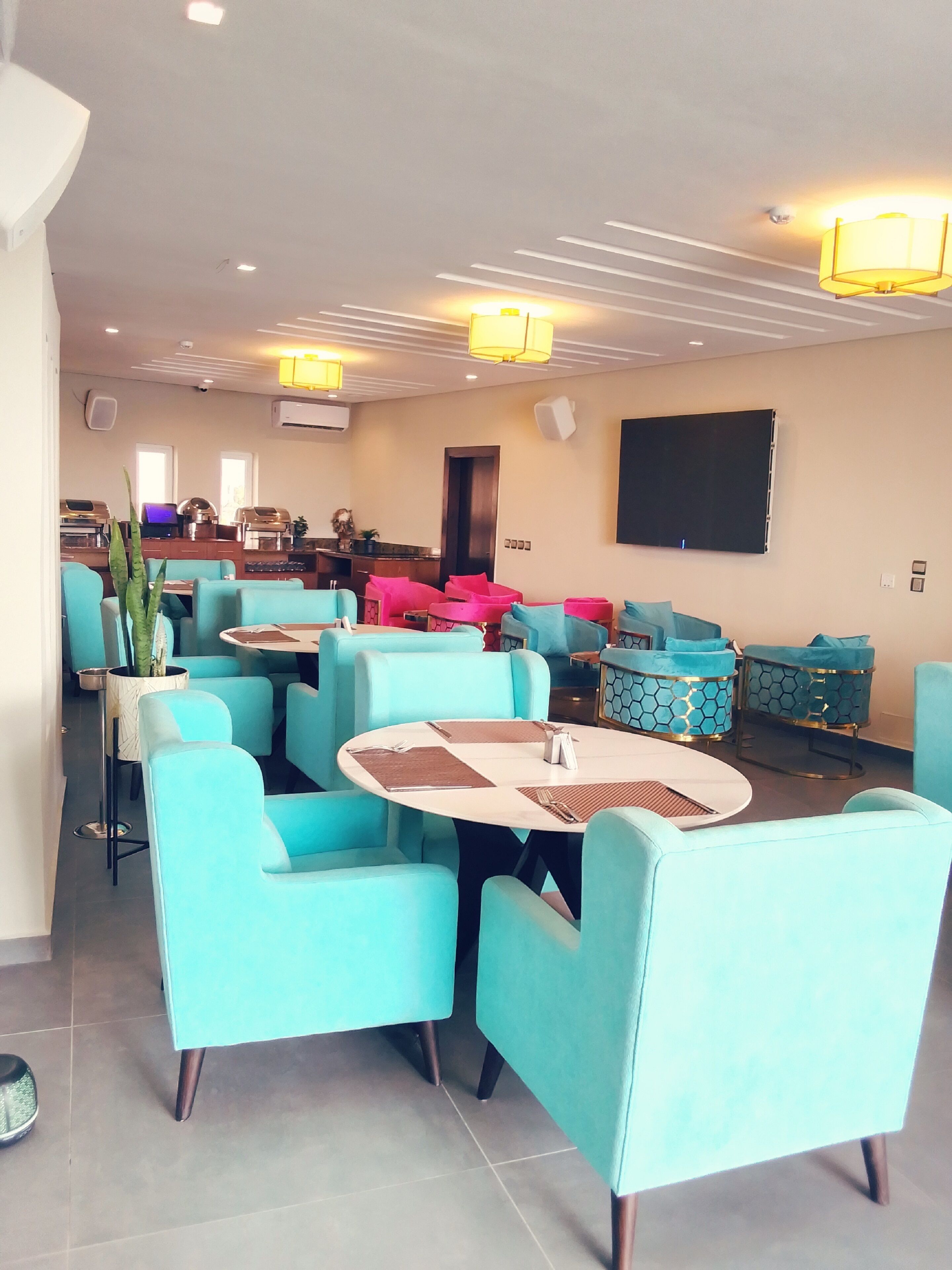 A spacious indoor area with teal and pink seating, a large flat-screen TV, and a kitchen area in the background.