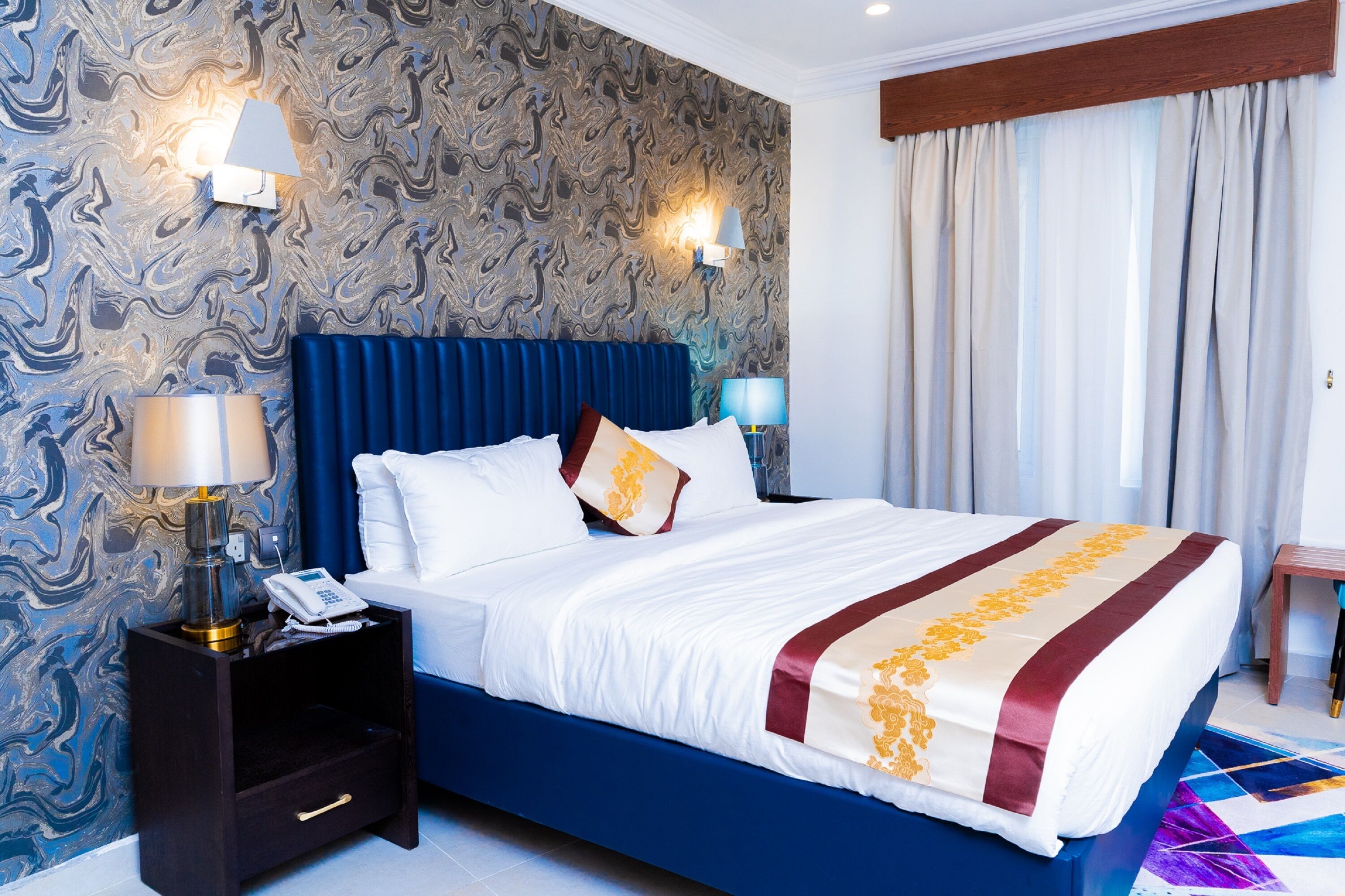A hotel room with a marble-patterned wall, a blue headboard, a white bed with a red and gold striped bedspread, two bedside lamps, and a nightstand with a phone and lamp.
