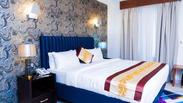 A hotel room with a marble-patterned wall, a blue headboard, a white bed with a red and gold striped bedspread, two bedside lamps, and a nightstand with a phone and lamp.