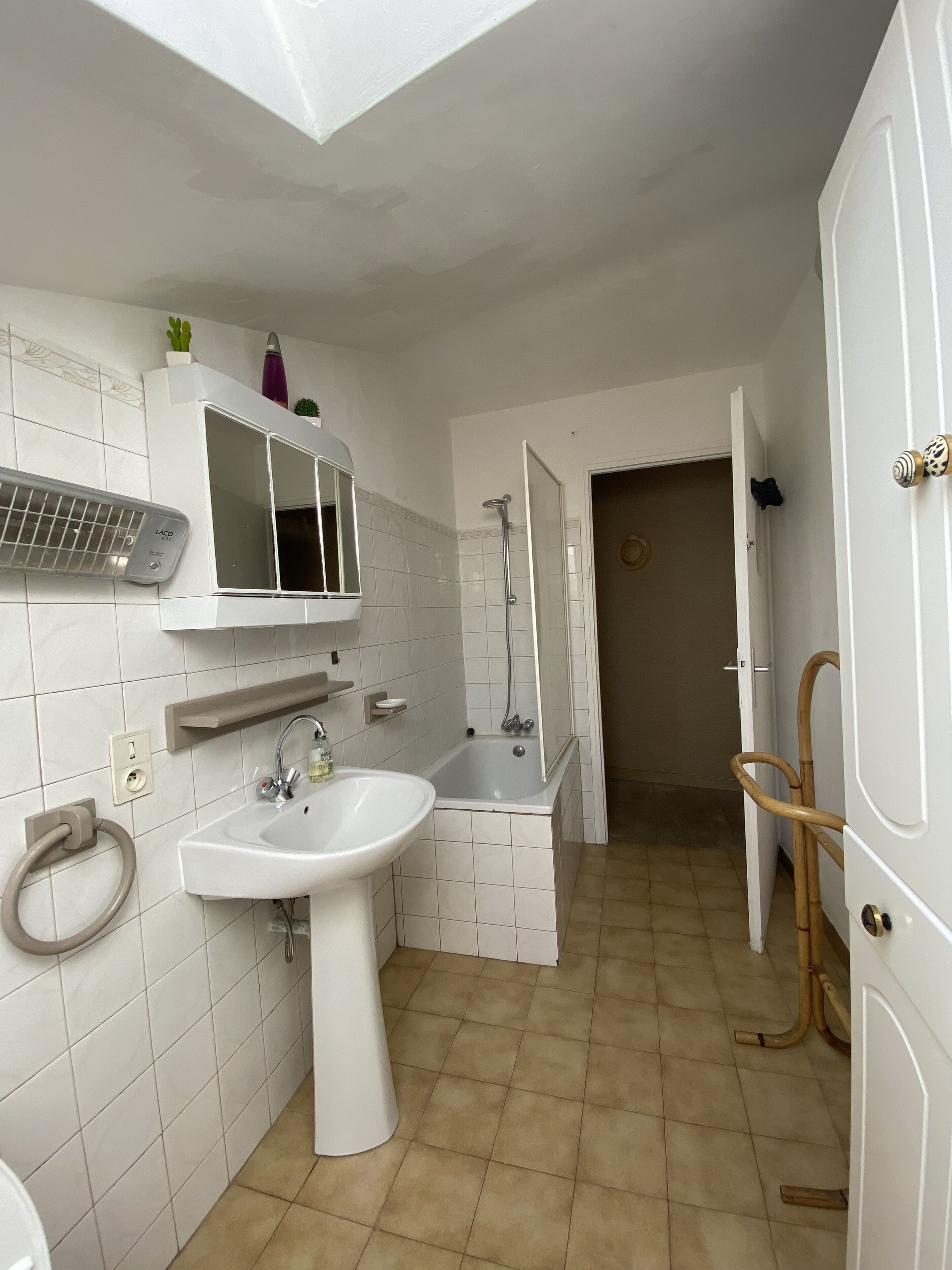 A bathroom with a white sink, mirror, and shower. There is a towel rack and a small shelf with toiletries. The floor is tiled, and the walls are white.