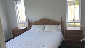 A bedroom with a wooden bed, white bedding, two nightstands, and windows with a view of a fence.
