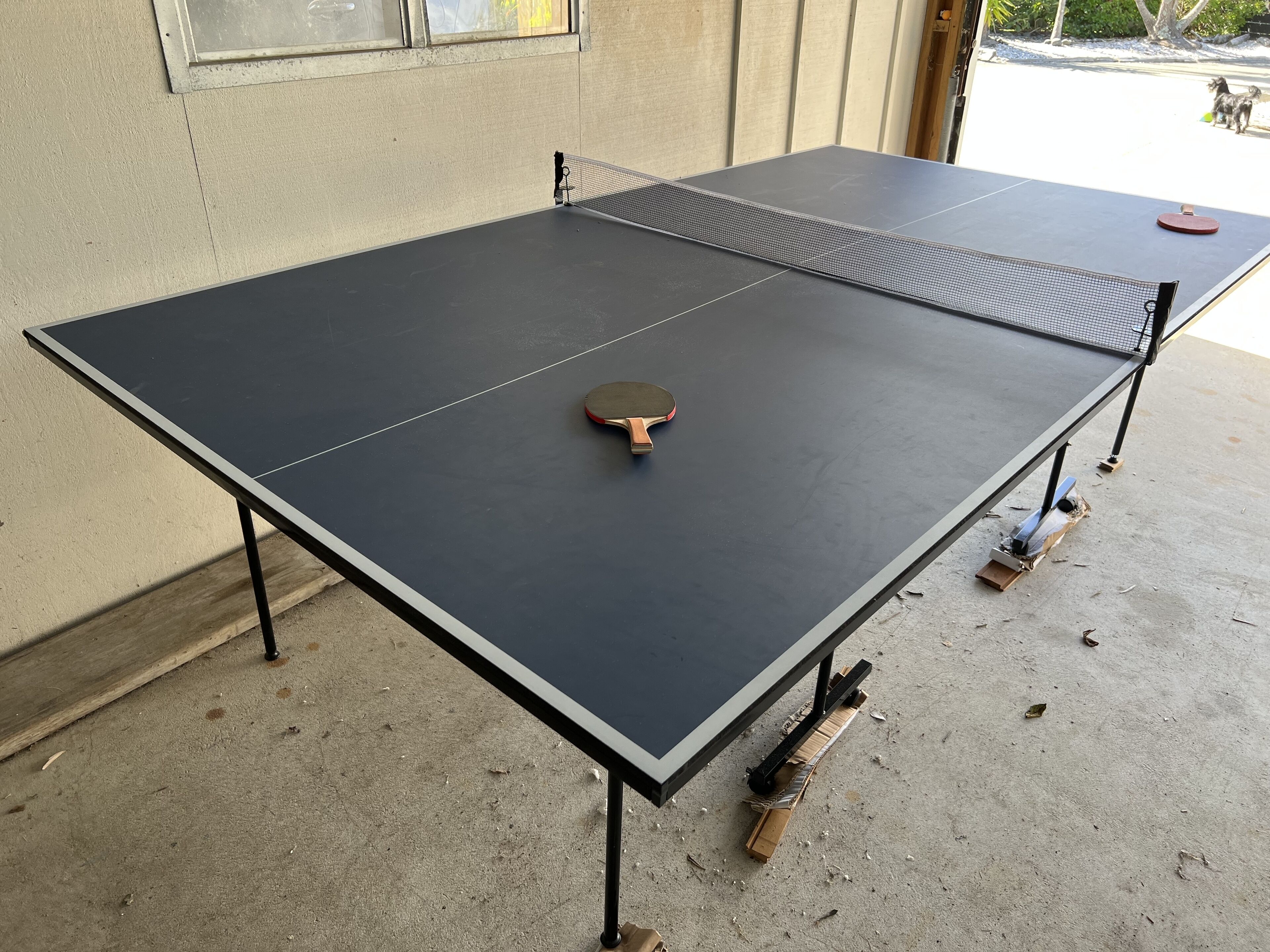 Une table de ping-pong avec une raquette, installée à l’extérieur sur un sol en béton, avec un pied de table en bois visible.