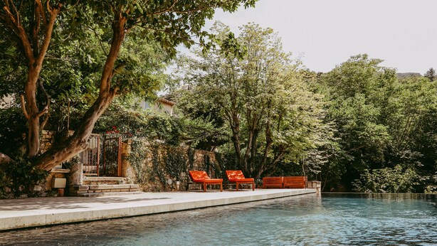 A swimming pool with a wooden deck and red lounge chairs, surrounded by lush greenery and trees.