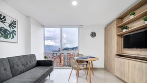 Un soggiorno moderno con un divano grigio, un tavolo da pranzo con sedie, una TV su una mensola in legno e un'ampia finestra con vista sulla città.