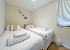 A bedroom with two beds, each with white linens and striped pillows. There are towels on the beds, and a window with blinds on the right. - 4 bedroom accommodation in Carnforth (Carnforth)