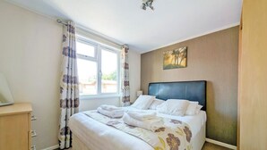 A neatly made bed with white linens and floral-patterned bedding, a wooden dresser with drawers, a window with curtains, and a painting on the wall.