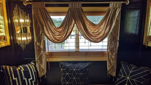 Une chambre avec un lit, une fenêtre avec des rideaux, un luminaire fixé au mur et un coussin à motifs.