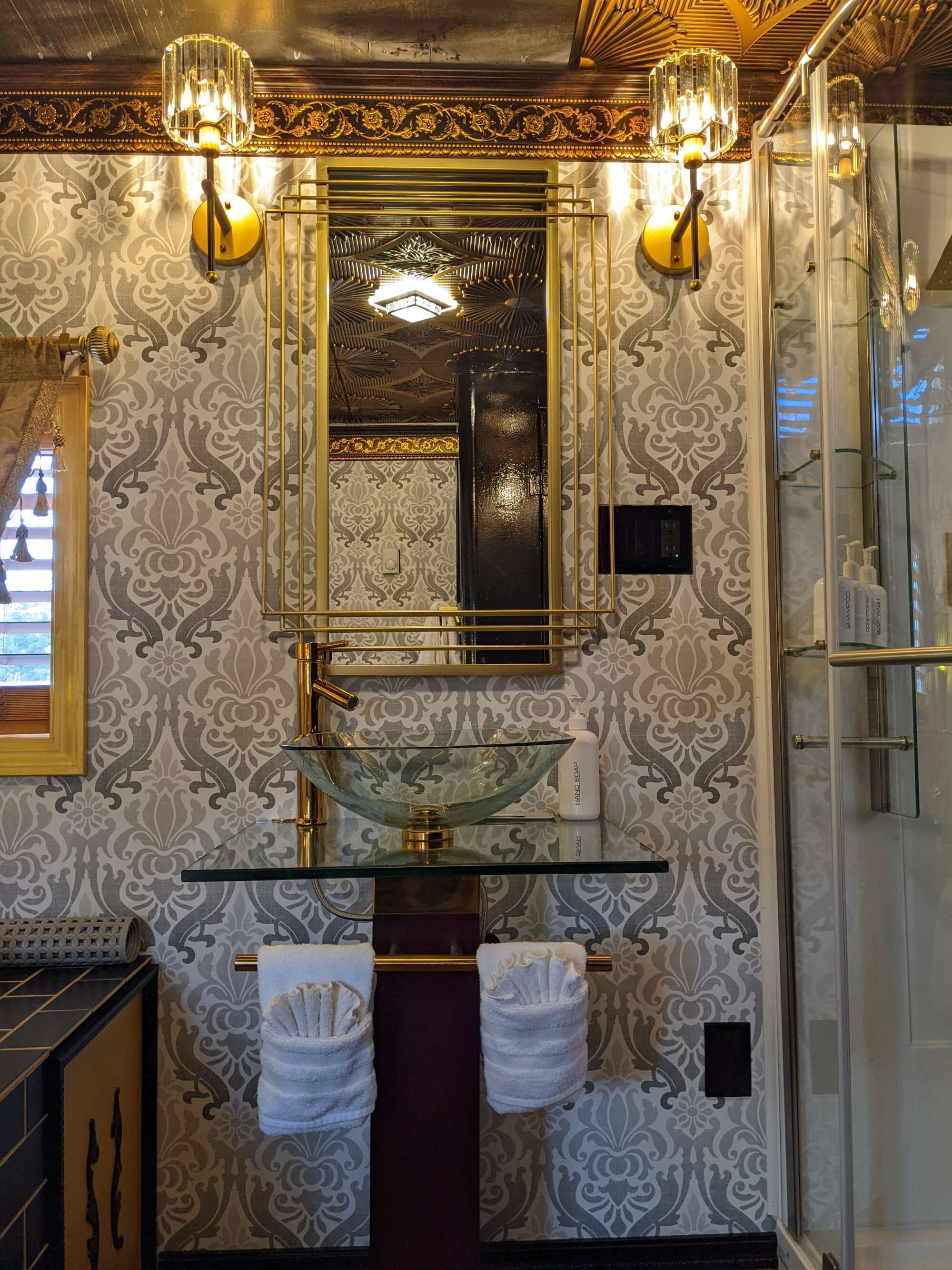 A bathroom with a glass bowl sink, two wall-mounted lights, a mirror, and patterned wallpaper.