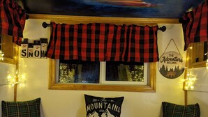 Une pièce chaleureuse avec un lambrequin à carreaux rouges et noirs au-dessus d’une fenêtre, une enseigne sur laquelle on peut lire « L’aventure commence », et un coussin orné du texte « Les montagnes nous appellent ».