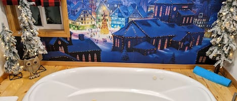 Ein Badezimmer mit einer großen weißen Badewanne, Holzboden und einem stimmungsvollen Wandgemälde eines verschneiten Dorfes.