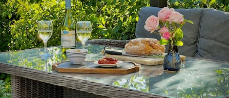 Una mesa de mimbre con superficie de vidrio, un pan, un cuenco de aceitunas, una botella de vino blanco y un jarrón con rosas rosadas.