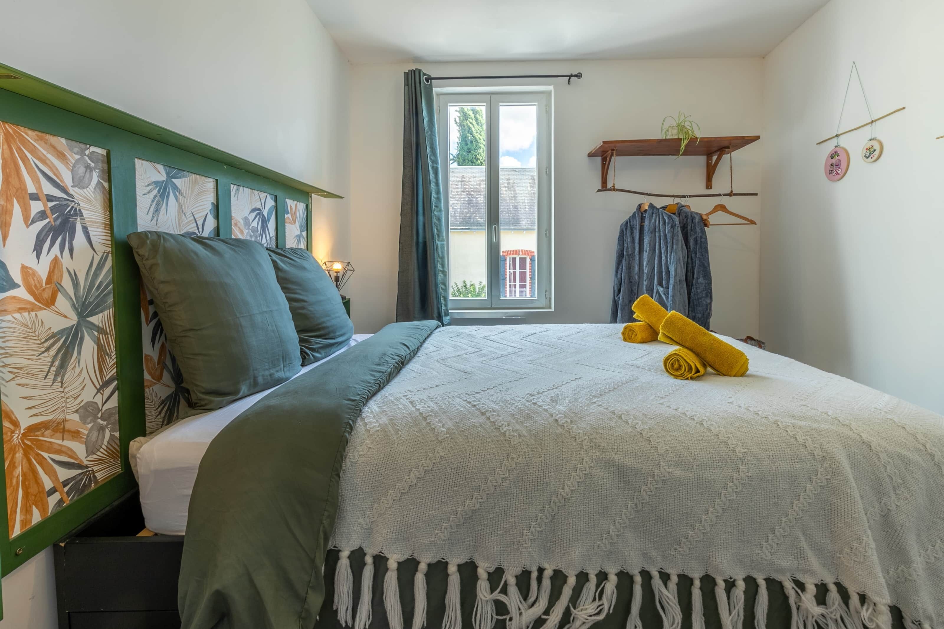 A bedroom with a bed, a window with curtains, a hanging plant, and a shelf with clothes and decorative items.