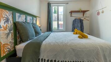 A bedroom with a bed, a window with curtains, a hanging plant, and a shelf with clothes and decorative items.