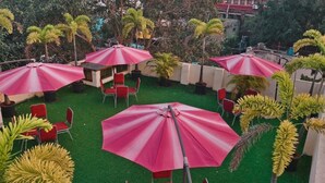An outdoor seating area with red chairs and pink umbrellas, surrounded by greenery and palm trees. - HOTEL ROYAL PARK (Chennai)