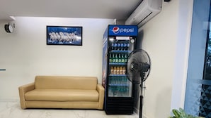 A room with a beige sofa, a wall-mounted clock, a Pepsi refrigerator, a standing fan, and a potted plant. - HOTEL ROYAL PARK (Chennai)
