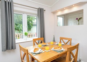 Un coin repas avec une table en bois, quatre chaises, des assiettes contenant des croissants et du jus d’orange, une fenêtre avec des rideaux et un radiateur.