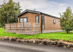 A single-story wooden cabin with a balcony, a stone retaining wall, and a grassy yard.