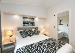A modern bedroom with a large bed, two bedside tables with lamps, a headboard with a cityscape artwork, and a bathroom visible through an open door.