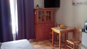 A small, compact room with a bed, a wooden cabinet, a table with a chair, and a television mounted on the wall.