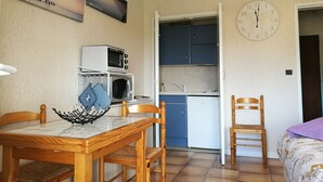 A compact kitchen with a microwave, a small refrigerator, and cabinets. There is a dining table with chairs and a clock on the wall.