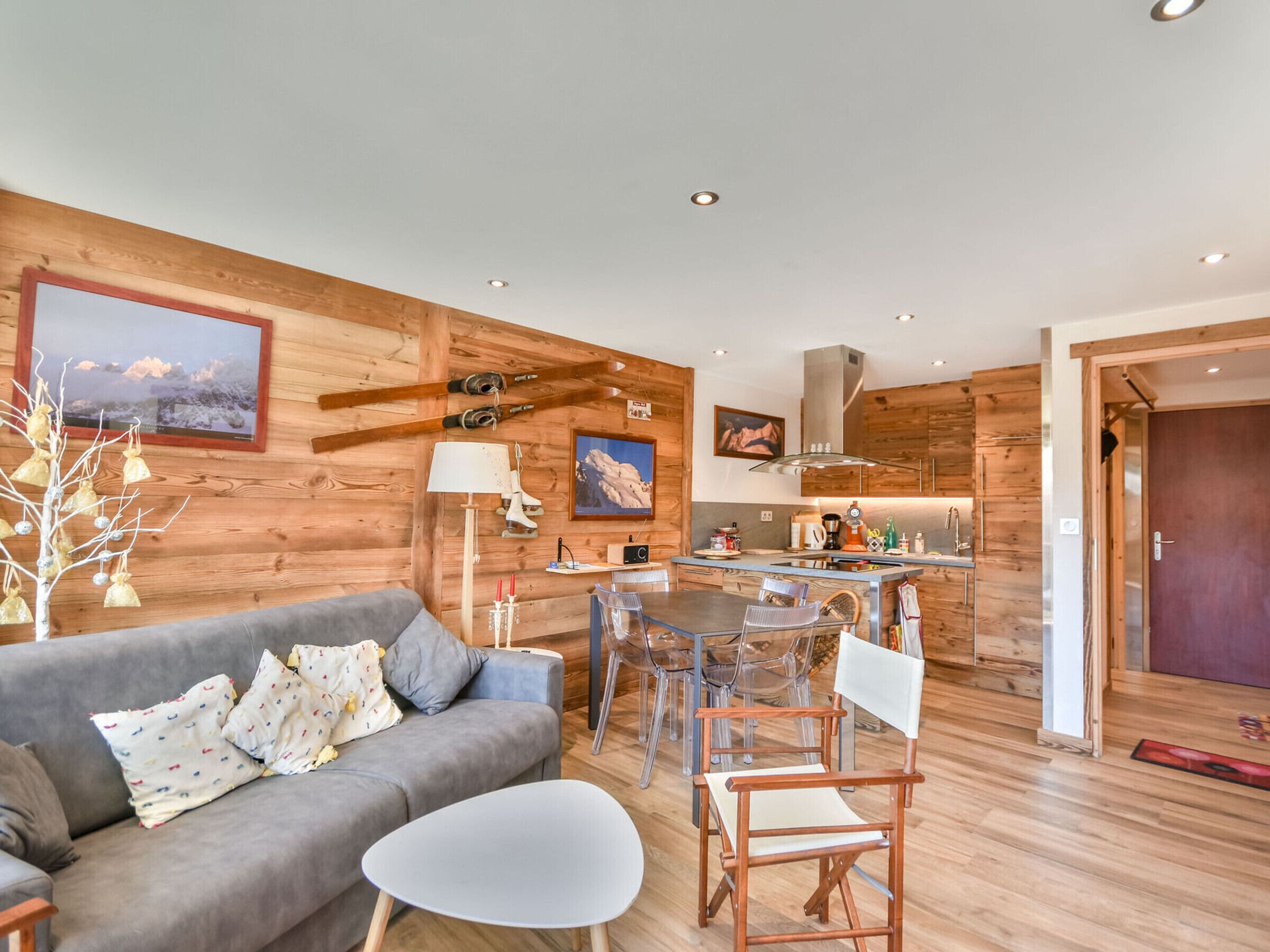 Ein modernes Wohnzimmer mit einem grauen Sofa, einem kleinen weißen Tisch und klaren Stühlen. Im Hintergrund ist eine Holz-Küche mit Herd und Spüle zu sehen.